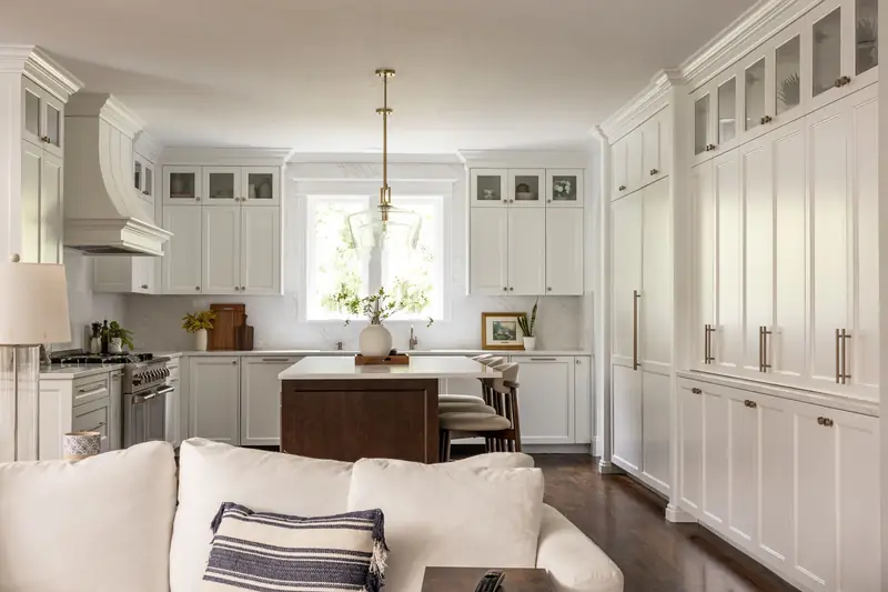 Open-concept view from the living room into the white kitchen, with a wall of tall built-in cabinets and brass hardware
