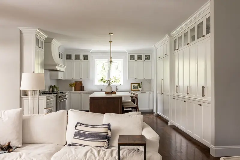 White kitchen seen from the family room, featuring a curved painted range hood, marble-look slab backsplash and glass-front upper cabinets