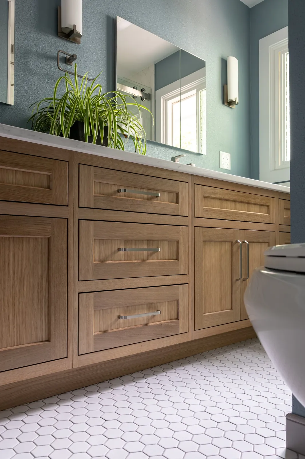 The finished bathroom: soft blue walls, a warm wood double vanity with quartz top, a wide mirror and white hexagon tile floor