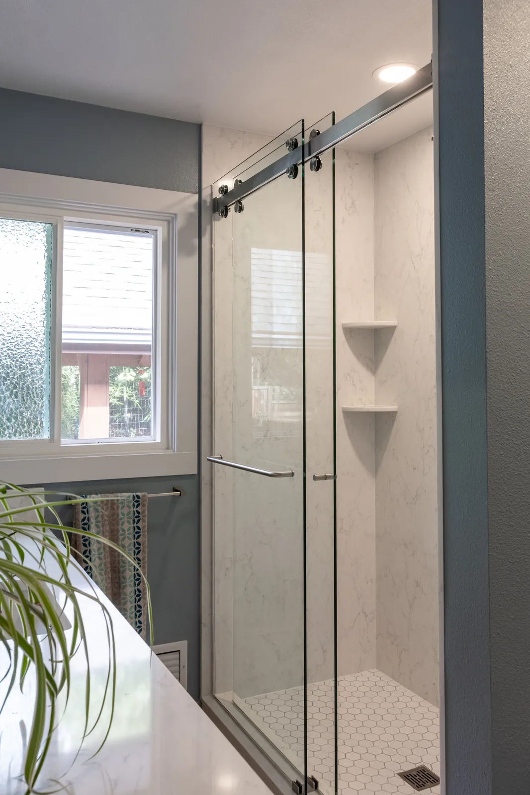 The same alcove after the tub-to-shower conversion: a walk-in shower with quartz slab walls, hexagon tile floor and a frameless sliding glass door