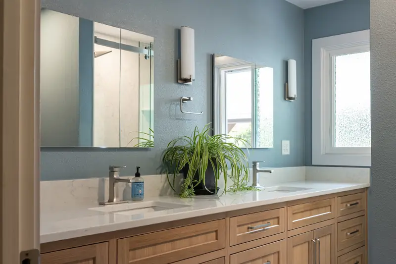 Wide view of the guest bath: wood vanity under a long mirror, soft blue walls and twin windows
