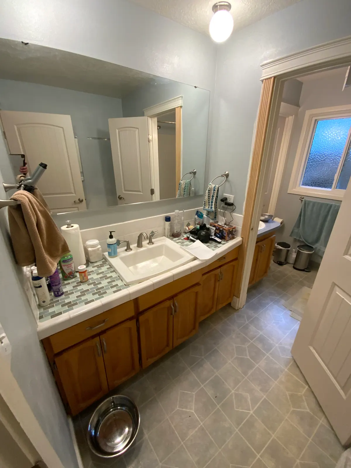 The original Jack-and-Jill bathroom before remodeling: a tile-topped oak vanity on one side of a partition wall, with a second vanity visible through the doorway beyond
