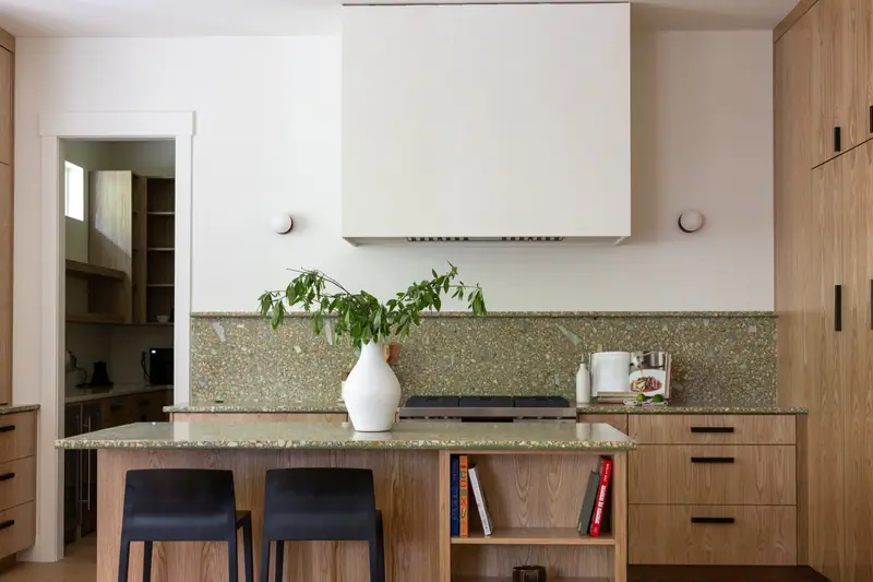 The plaster hood over the island cooktop, framed by full-height white oak cabinetry and the green terrazzo-look quartz backsplash
