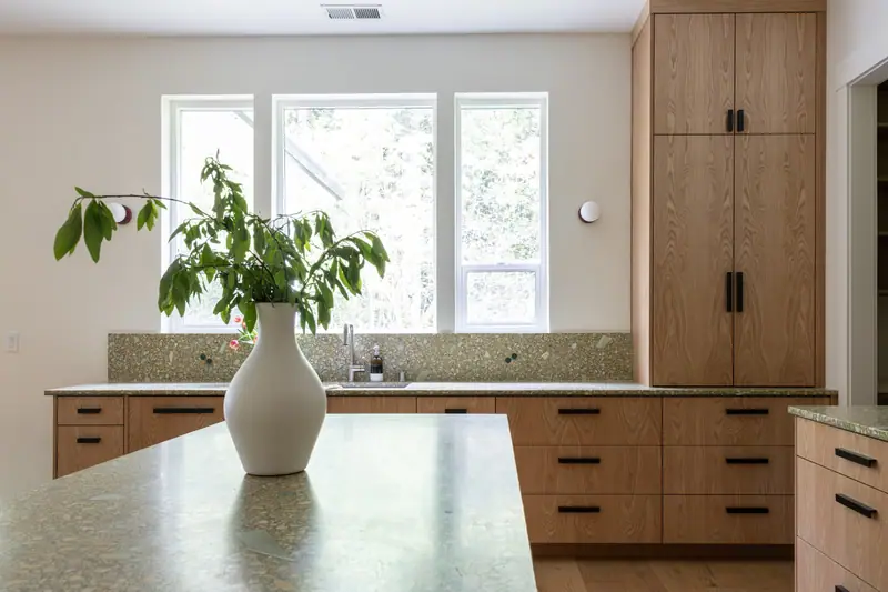 The sink wall head-on, with three tall windows looking into trees, quartz counters and backsplash, and full-height white oak cabinetry to the right