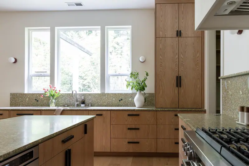 Daylight across the quartz counters from the window wall, with the stainless pro-style range and a built-in oven set into oak cabinetry