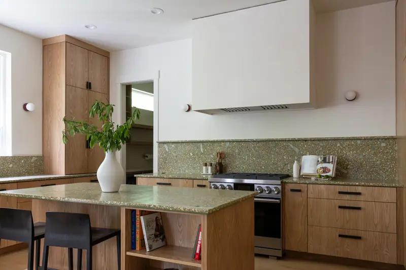 The island and cooking wall together, with the stainless range under the plaster hood and cookbooks in the open end of the island