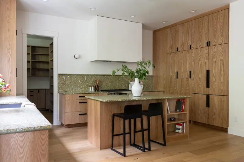 The kitchen from the hall doorway: pantry entry at the left, oak island with two black stools, and a full-height run of white oak cabinetry