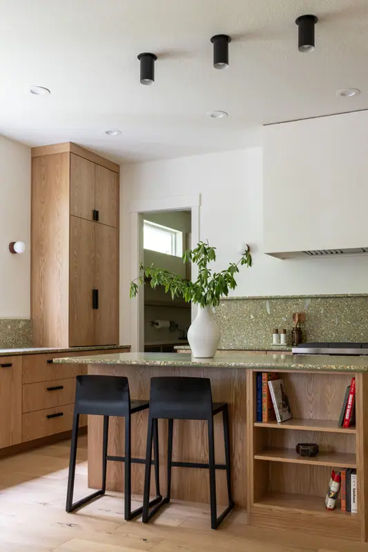 The island with its bookshelf end and black stools, with a clerestory window and the plaster hood beyond