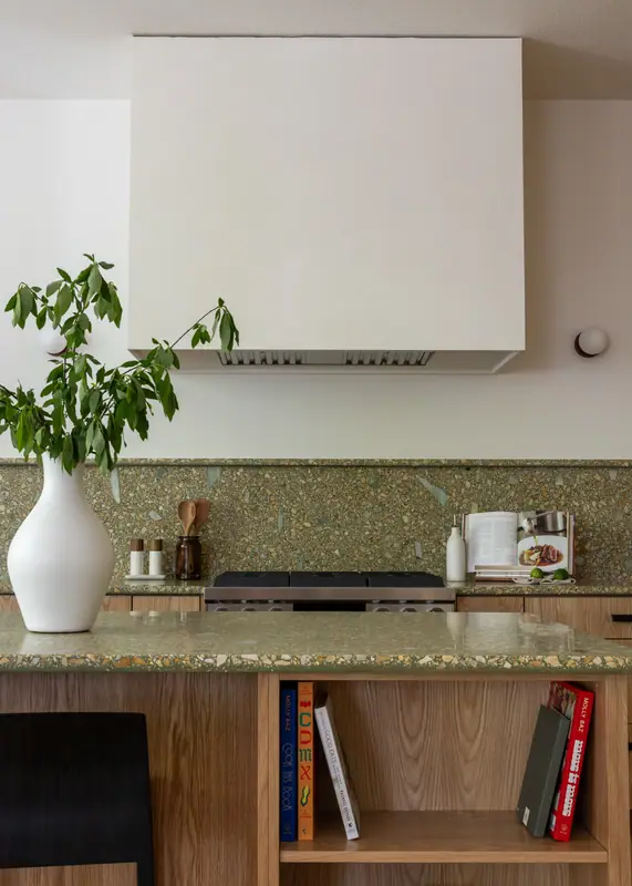 A vase on the quartz island top, with the cooktop and plaster hood behind and cookbooks in the island shelf