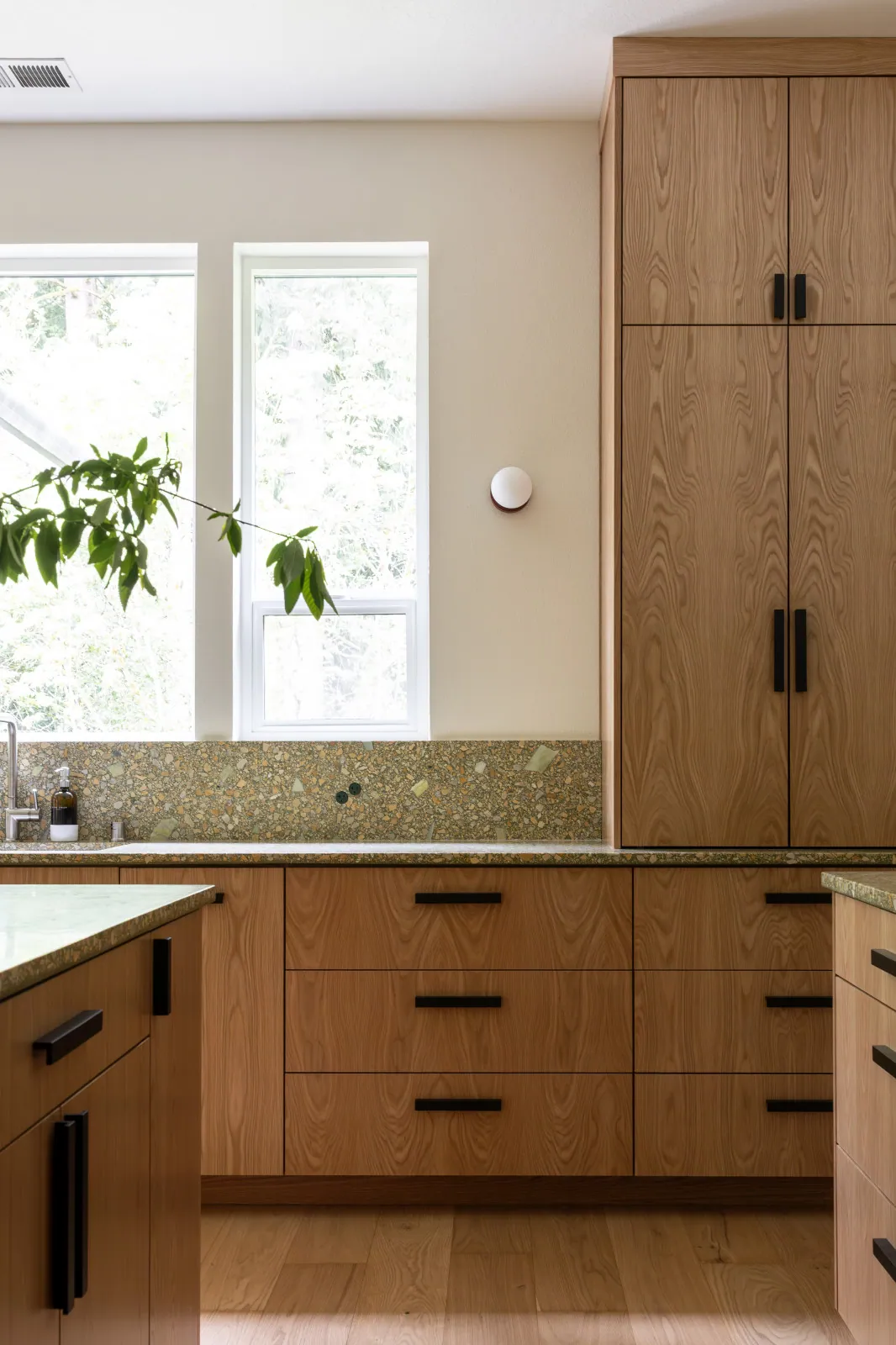 The tall plain-sliced white oak cabinet that replaced the original black open-shelving unit at the end of the sink run, with the terrazzo-look quartz counter and full-height backsplash alongside