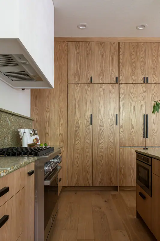 The range under the plaster hood with its exposed stainless liner, flanked by panel-clad white oak appliance columns and a built-in oven