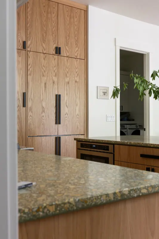 The panel-clad refrigerator columns in plain-sliced white oak with long black pulls, beside the built-in oven and the quartz island