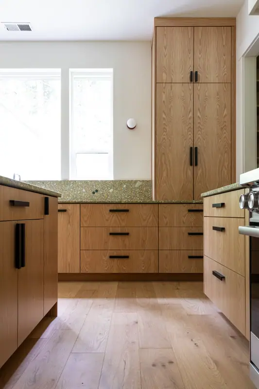 The base cabinets of the sink run with their long black pulls, the range at the right edge and wide-plank oak floors