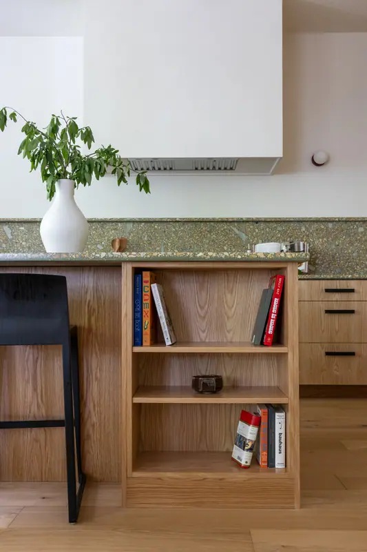 The open oak bookshelf end of the island holding cookbooks, with a black stool and the plaster hood behind