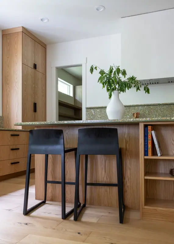 The island with two black counter stools and its bookshelf end, in front of the oak cabinetry and quartz counters
