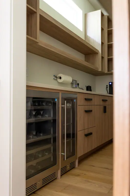 The glass-front wine refrigerator built into the pantry counter, with the coffee station above and oak open shelving overhead