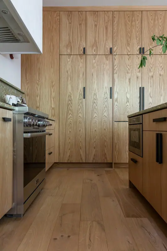 The appliance wall with the refrigerator concealed behind full-height white oak panels, the stainless range at the left and a built-in oven at the right