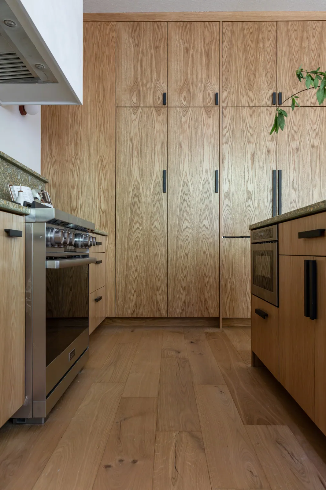 The same wall after remodeling: floor-to-ceiling plain-sliced white oak with the refrigerator hidden behind matching panels, the oven built into oak-fronted cabinetry and the stainless range left as the one visible appliance