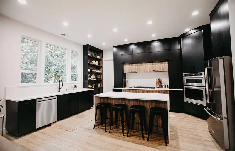 Before the remodel: the original dark kitchen seen from the dining end, with matte black cabinetry to the ceiling, black base cabinets under the windows, a black open-shelving unit and a wood-panelled island