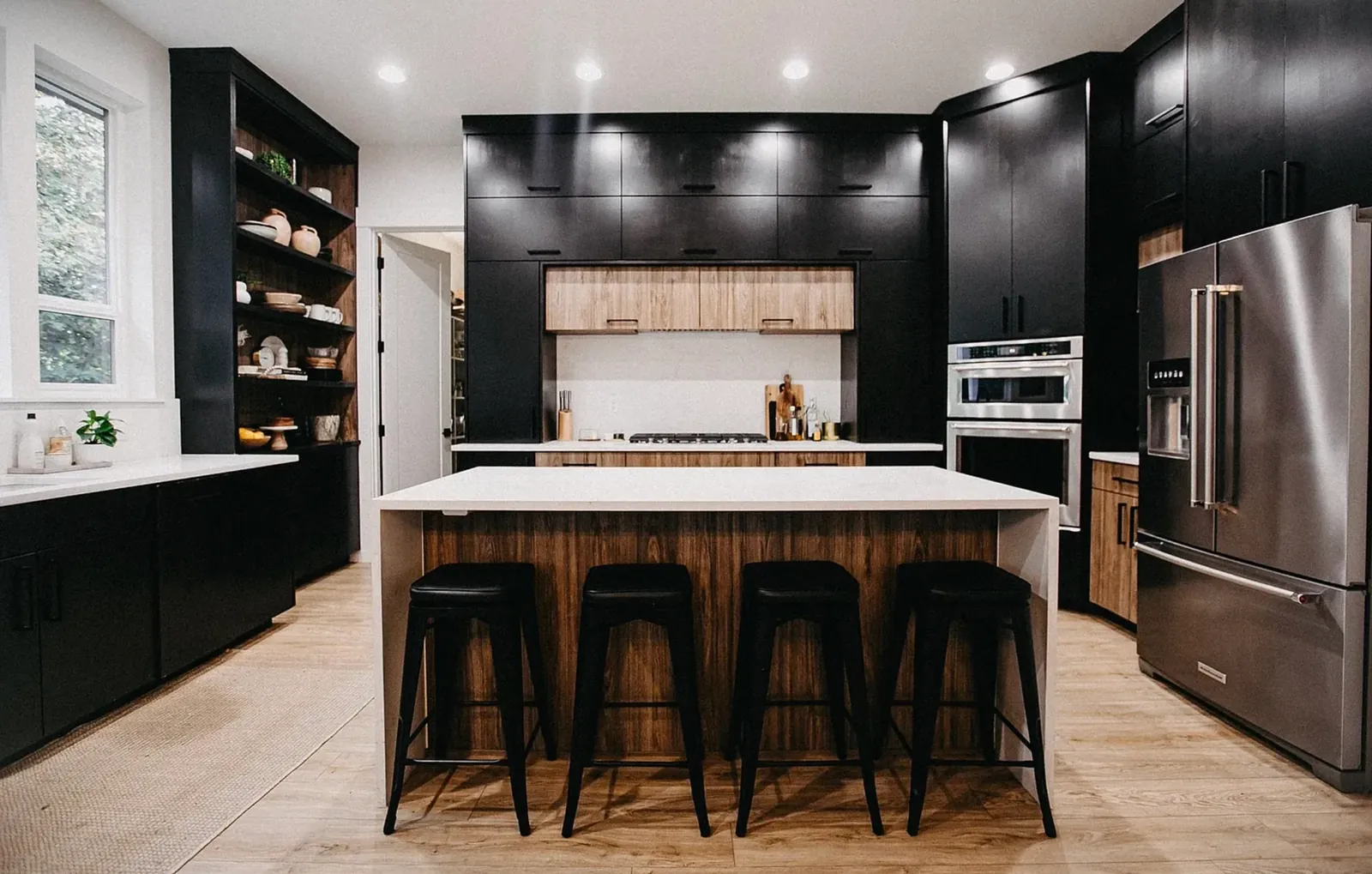 The original cooking wall head-on before remodeling, with black upper cabinets, a dark band above the cooktop and a wood-panelled island with a white quartz top