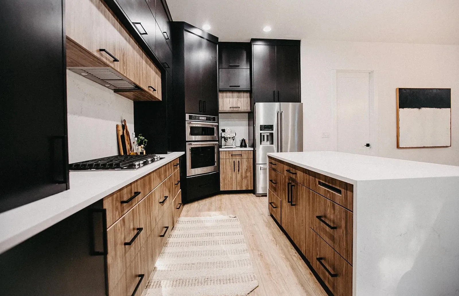 The original appliance wall before remodeling: a stainless oven stack and an exposed stainless French-door refrigerator breaking up the black cabinetry, with wood drawer banks and white quartz counters