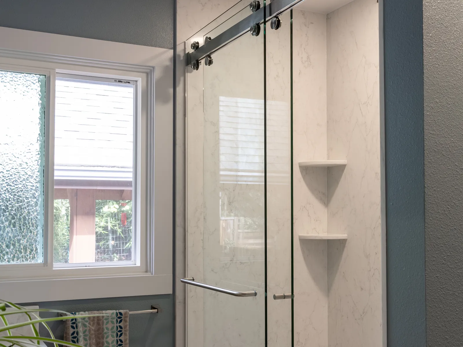 Walk-in shower with a frameless sliding glass door, white marble-look wall panels, corner shelves and a window beside it